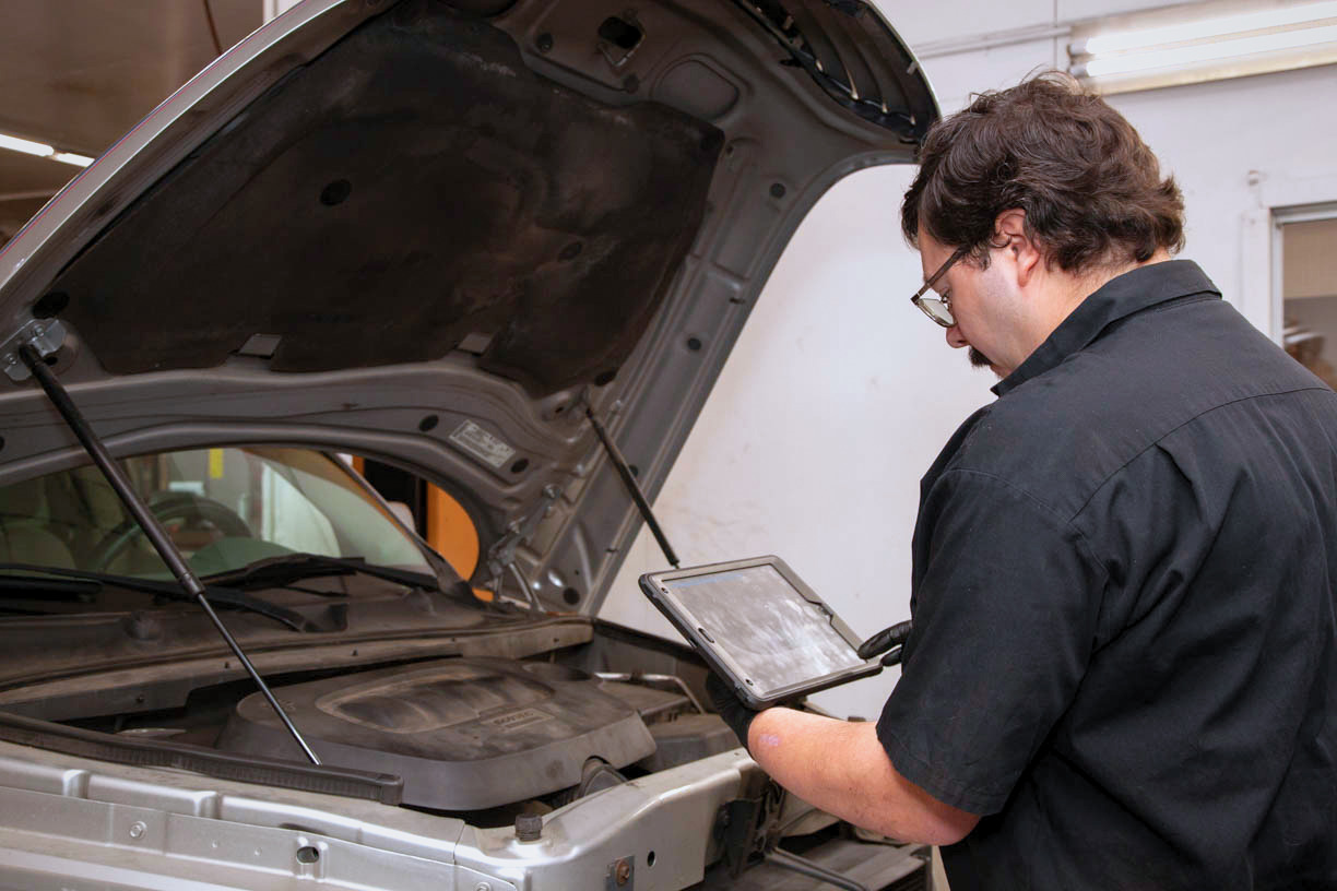 Technician using a diagnostic tablet beside a car with its hood open